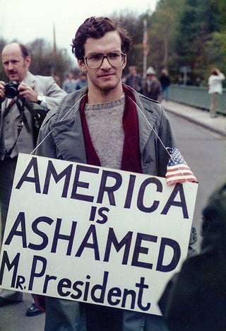 Protester holding "America is ashamed Mr. Presdent" placard over POTUS Ronald Reagan visiting Kolmeshöhe Cemetery, a German military cemetery containing Waffen-SS graves. Bitburg, West Germany. May 5, 1985 [500x732]
